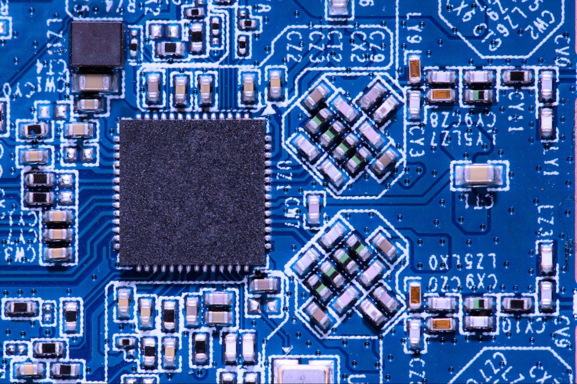 High-resolution macro photograph of a modern blue circuit board. It features a central QFP integrated circuit surrounded by precision-soldered surface-mount (SMD) resistors and capacitors.