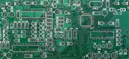 Detailed view of a bare green FR4 printed circuit board. The image highlights the copper pads, drill holes, and white silkscreen identifiers used for assembly.